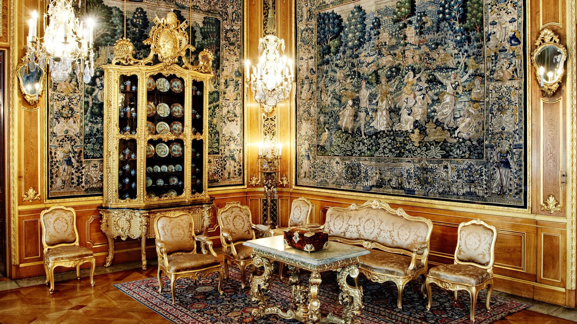 Ett rum med träpaneler med gulddetaljer och en sittgrupp med förgyllda möbler. I bakgrunden ett förgyllt skåp med porslinstallrikar inuti.