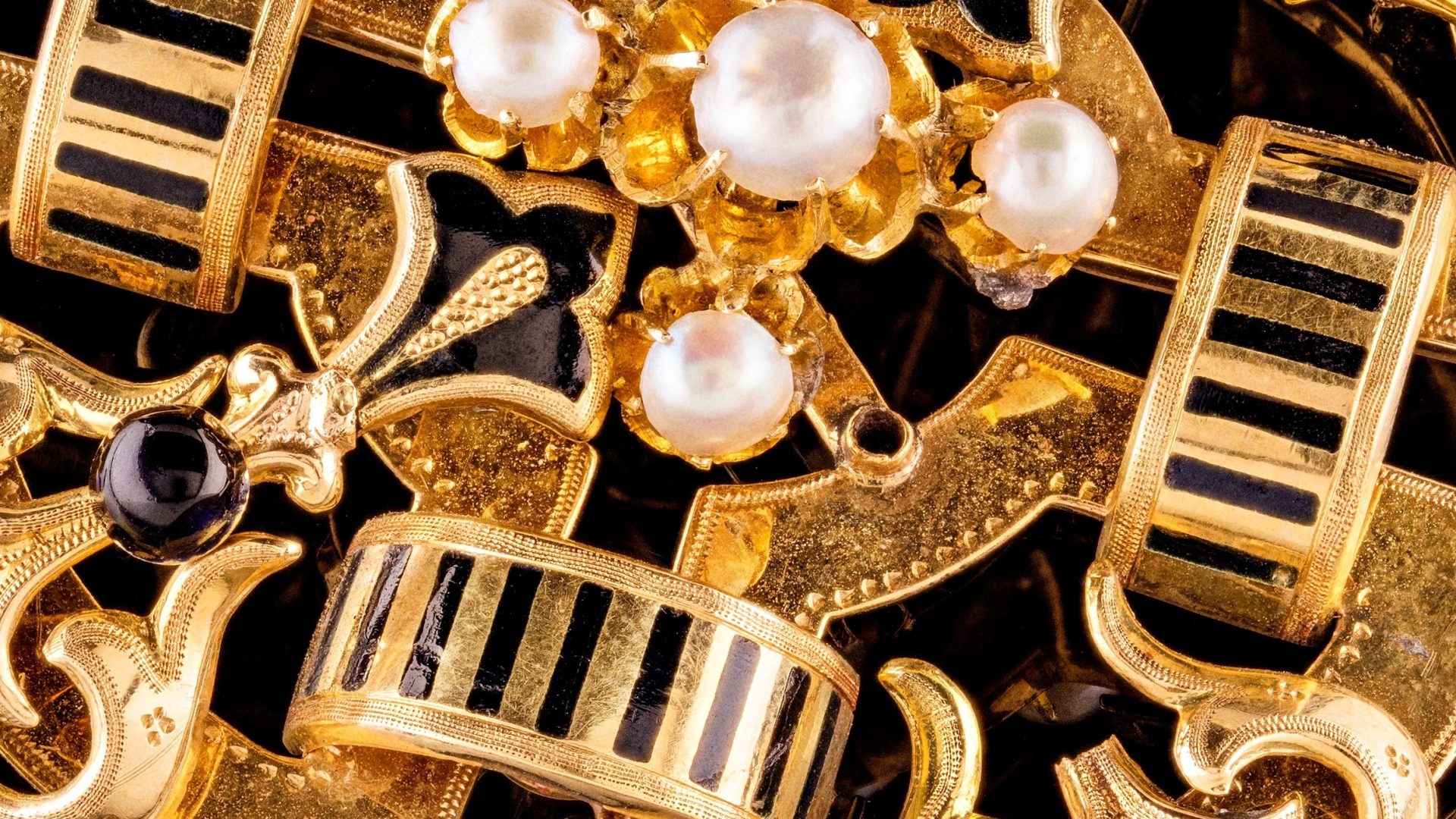 Detaljbild av ett smycke av guld med svart emalj och vita pärlor.