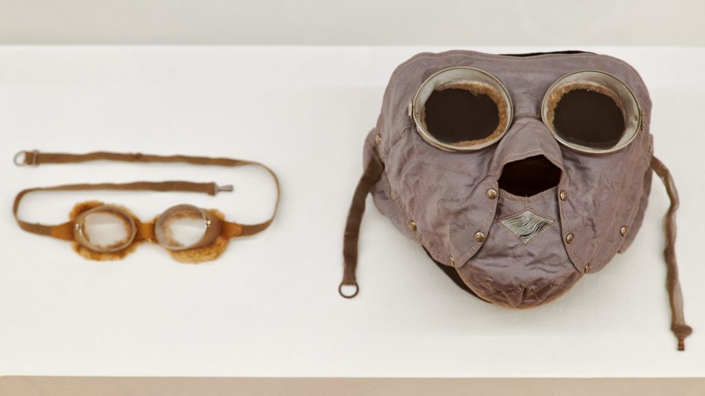 A pair of goggles and a fully covering face mask of grey fabric, used when driving an automobile in the early 1900s.