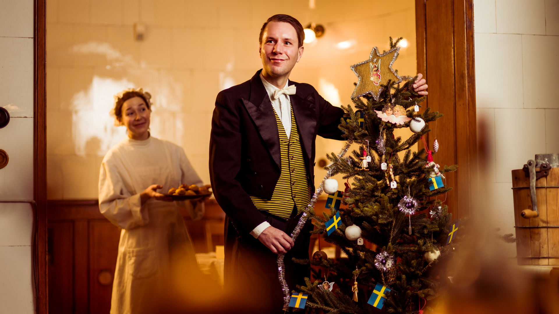 One person decorating a Christmas tree, and another holding a tray of saffron buns (lussebullar).