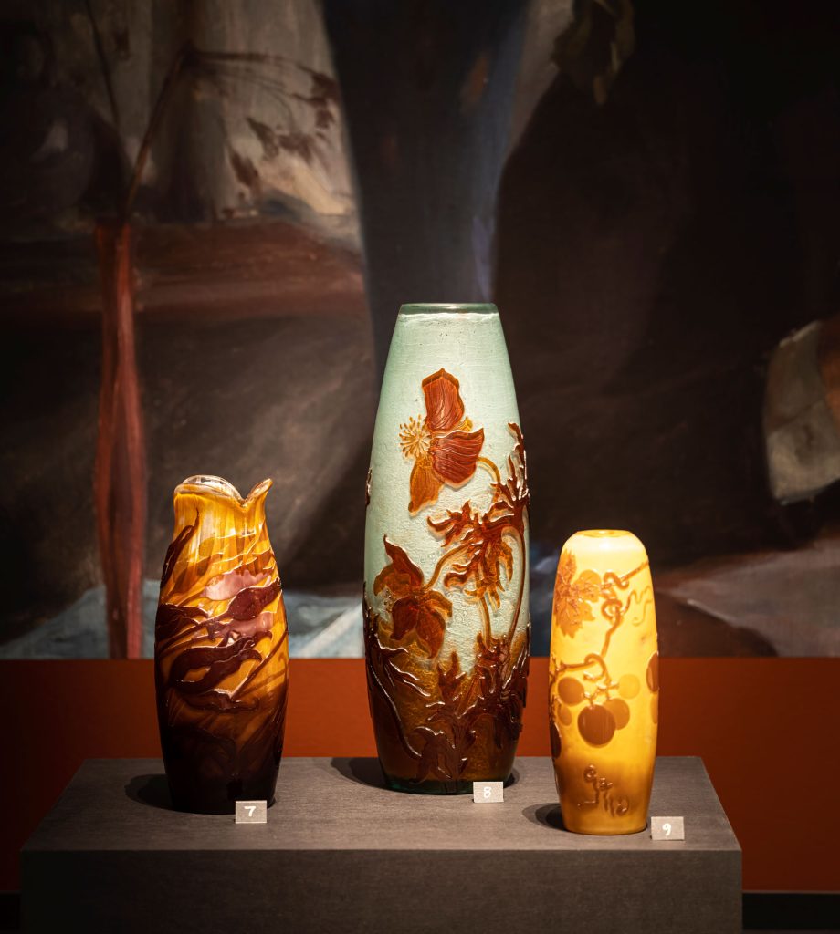 Tre vaser på ett podium. En vas med mönster föreställande blomknoppar i strömmande vatten, en annan vas med mönster som föreställer anemoner och en tredje med vindruvsdekor