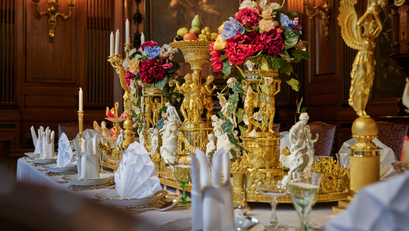 Ett uppdukat bord med bland annat kandelabrar, fint vikta servetter stående på vitt och guldigt porslin.