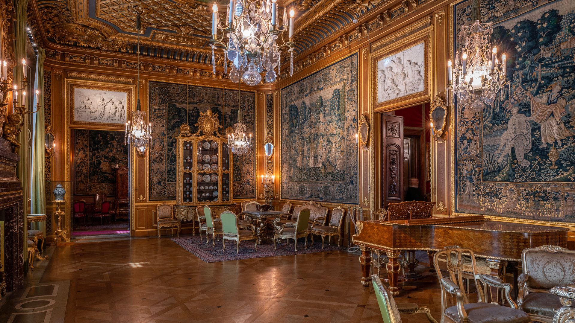 Ett stort rum med väggar i trä med gulddetaljer och stora tygtapeter med grönaktigt mönster. I mitten av bilden en flygel och en sittgrupp med förgyllda möbler.