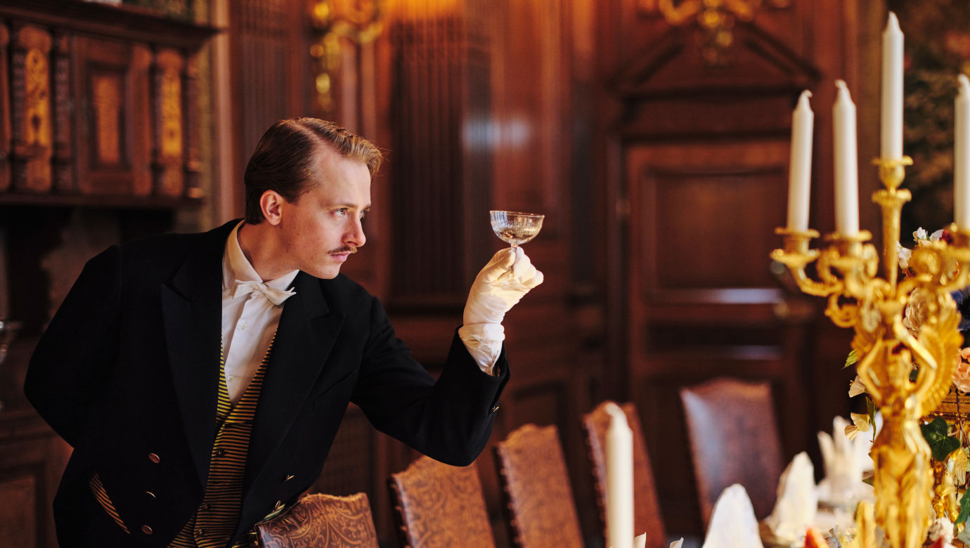 A person in a tail-coat stands next to a beautifully set dining table, holding a well-polished wineglass. 