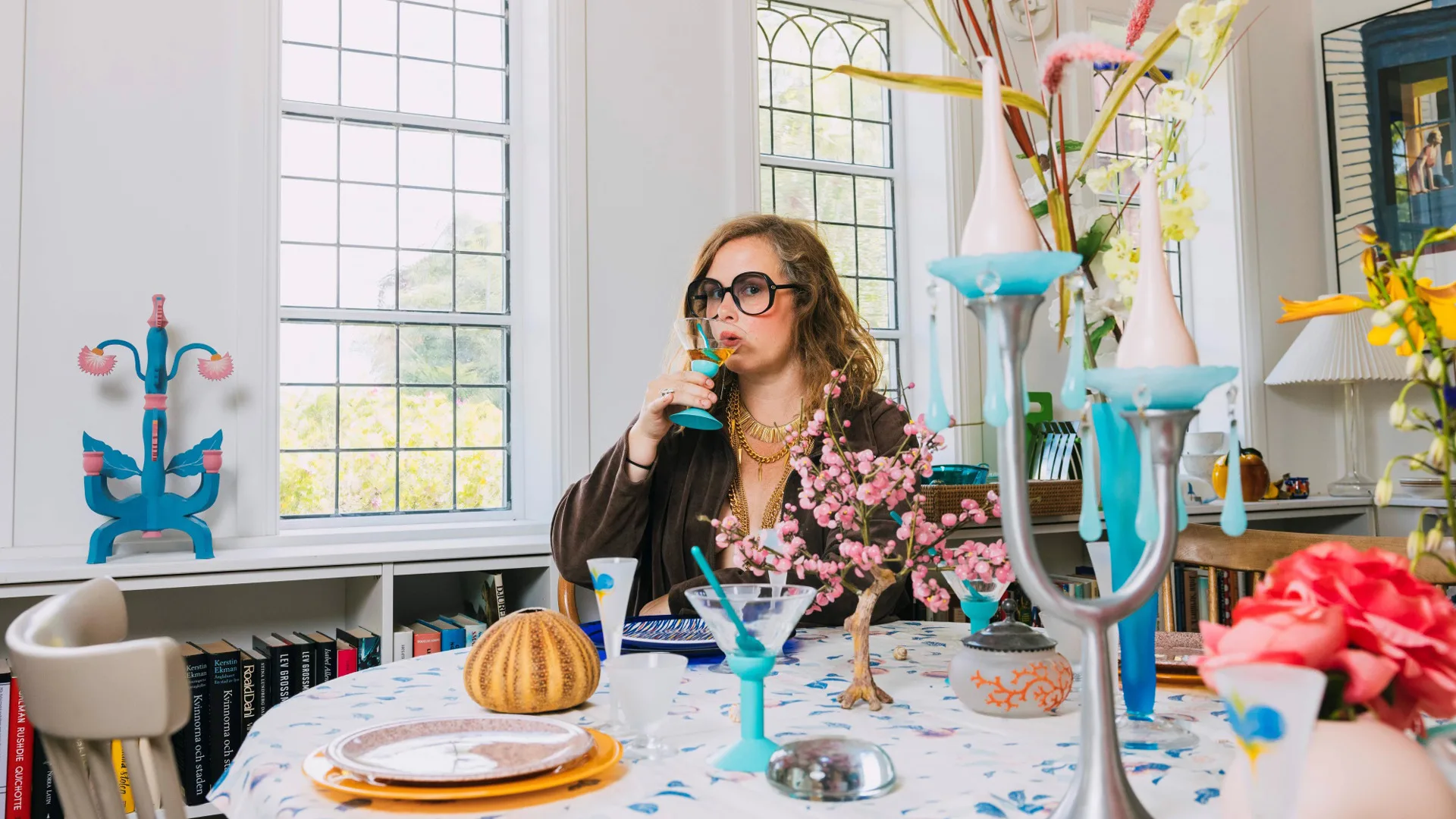 Hanna Hellquist sitter vid ett uppdukat bord med blommor och ljusstakar. Hanna dricker ur ett vinglas.