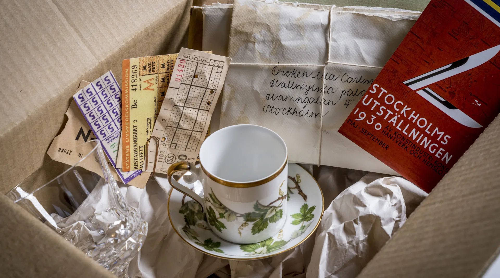 En flyttlåda med gamla saker som en kafffekopp med tillhörande fat, vinglas i kristall, en bunt med restaurangkort, en bunt med brev och kuvert