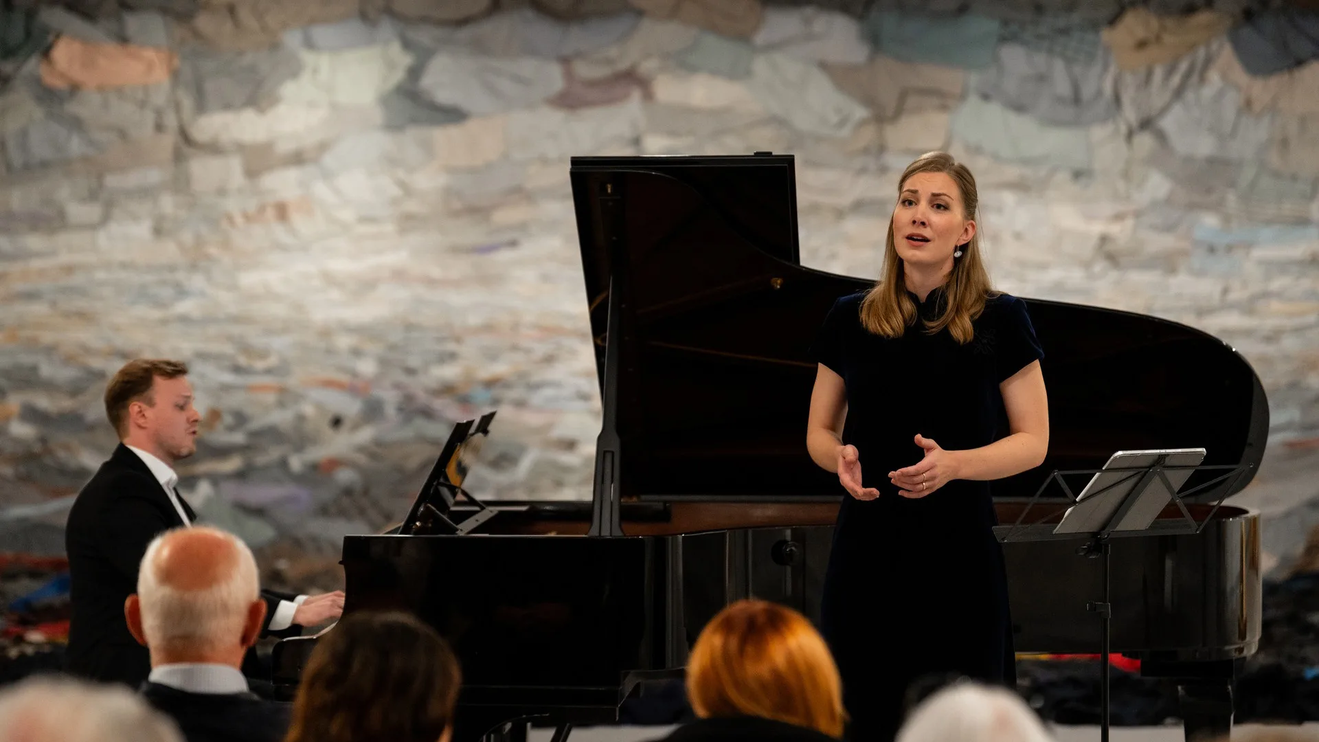 En kvinna i svart klänning sjunger framför ett piano. Pianot spelas av en man i kostym.