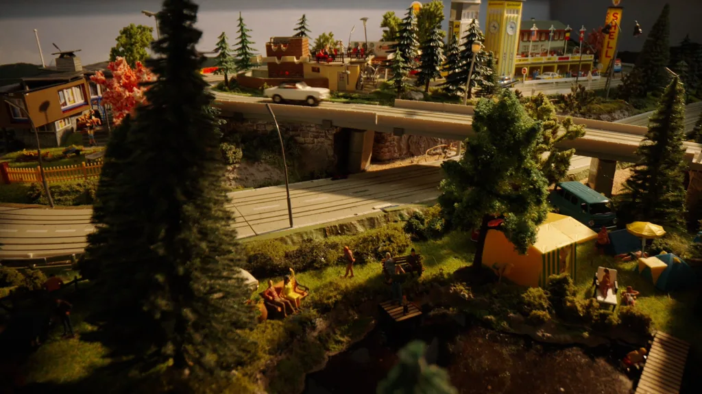 Detaljerad miniatyrmodell föreställande en småstad med campingplats i förgrunden. Scenen inkluderar tält, människor som campar, badar och umgås vid ett litet vatten, med vägar och broar som korsar landskapet. I bakgrunden syns byggnader, bilar, träd och ett Shell-bensinstationsskylt.