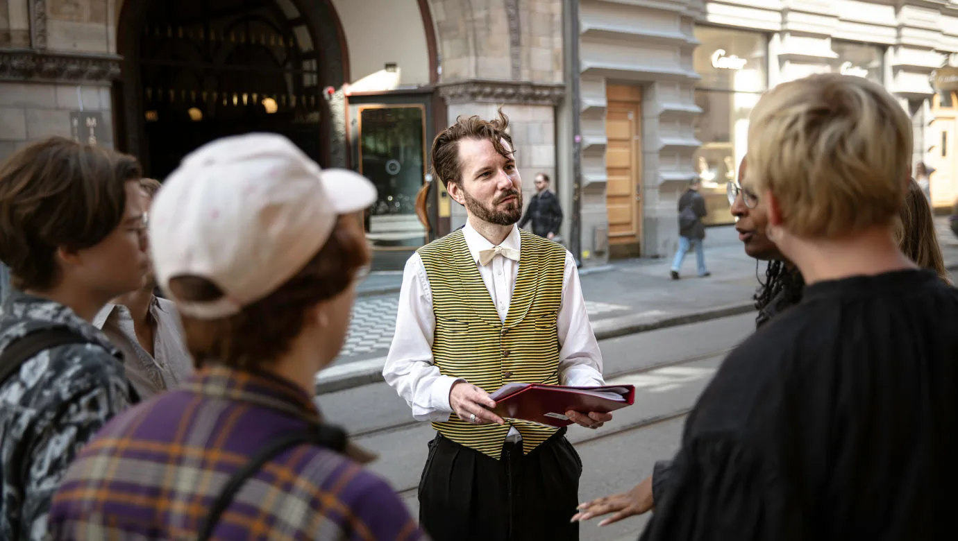 En guide i randig väst och fluga håller i en pärm och pratar med en grupp personer under en stadsvandring utanför en historisk byggnad i centrala Stockholm.