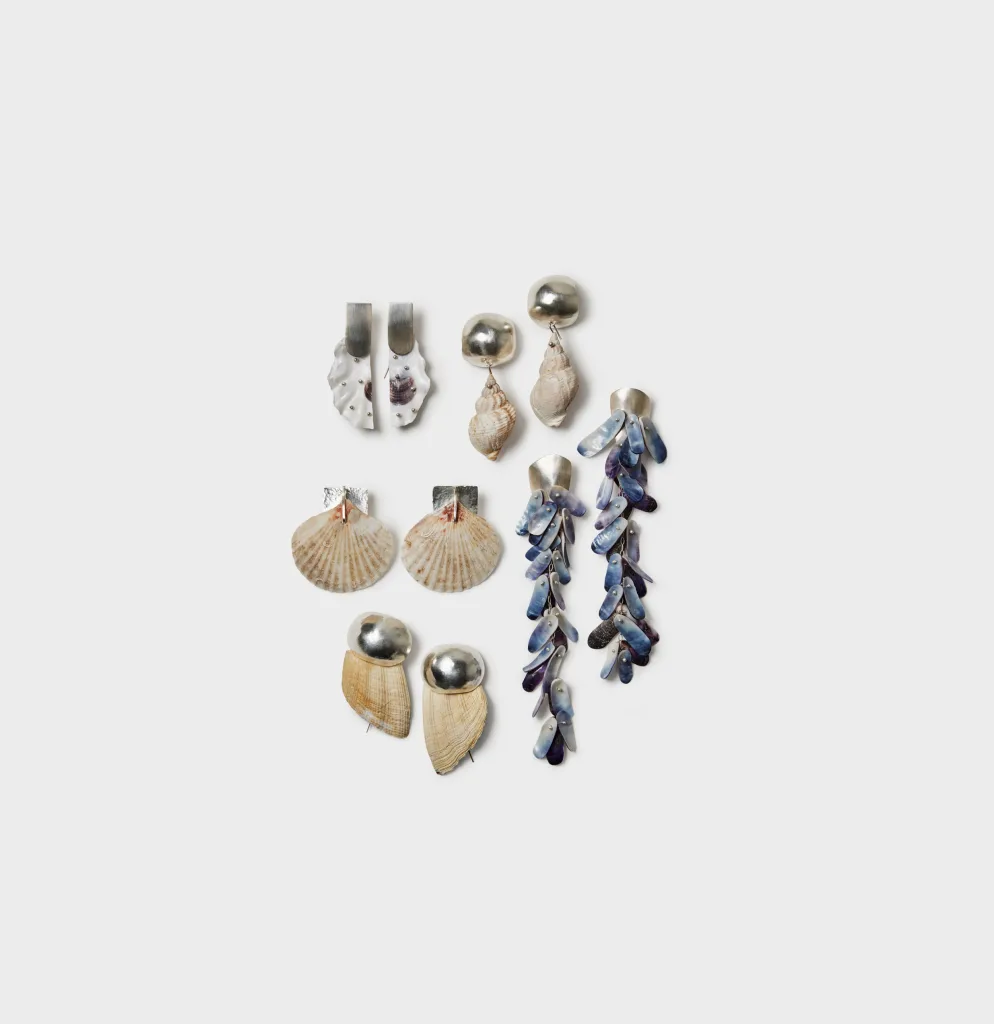 En samling örhängen arrangerade på vit bakgrund, tillverkade av snäckor, pärlemor och silver. Örhängena varierar i form och färg – från korta och skulpturala till långa, hängande modeller. Några är gjorda av flata kammusslor, andra av spiralformade snäckor, och ett par består av blåskimrande pärlemorliknande delar som hänger i flera nivåer.