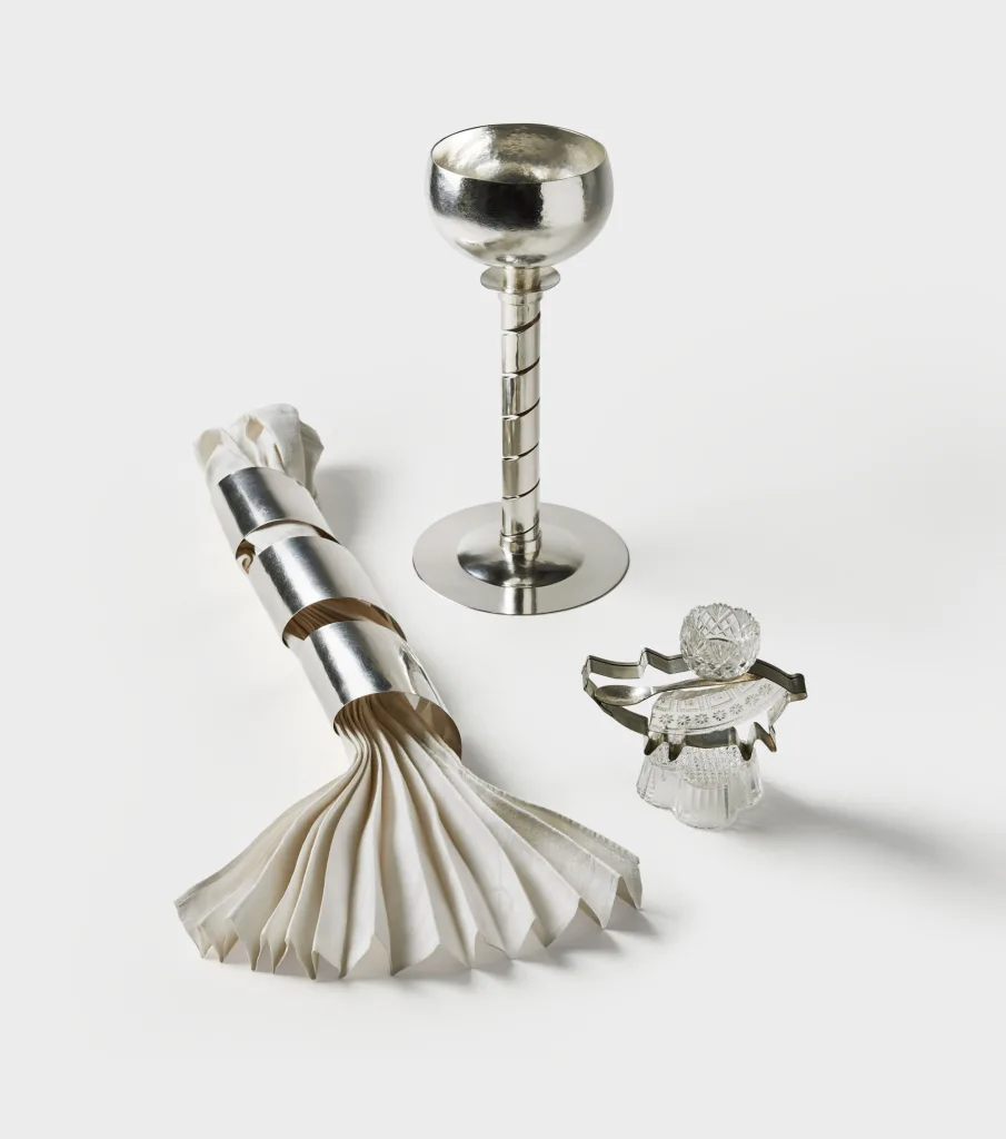 Ett stilleben mot vit bakgrund med tre eleganta objekt i metall och glas. Till vänster ligger en vikt tygservett omsluten av en bred, spiralformad servettring i silver. I mitten står en hög, silverglänsande bägare med en spiralformad fot. Till höger syns ett dekorativt föremål i glas och metall som liknar en liten figur med utsvängd kjol och kristallkula som huvud.