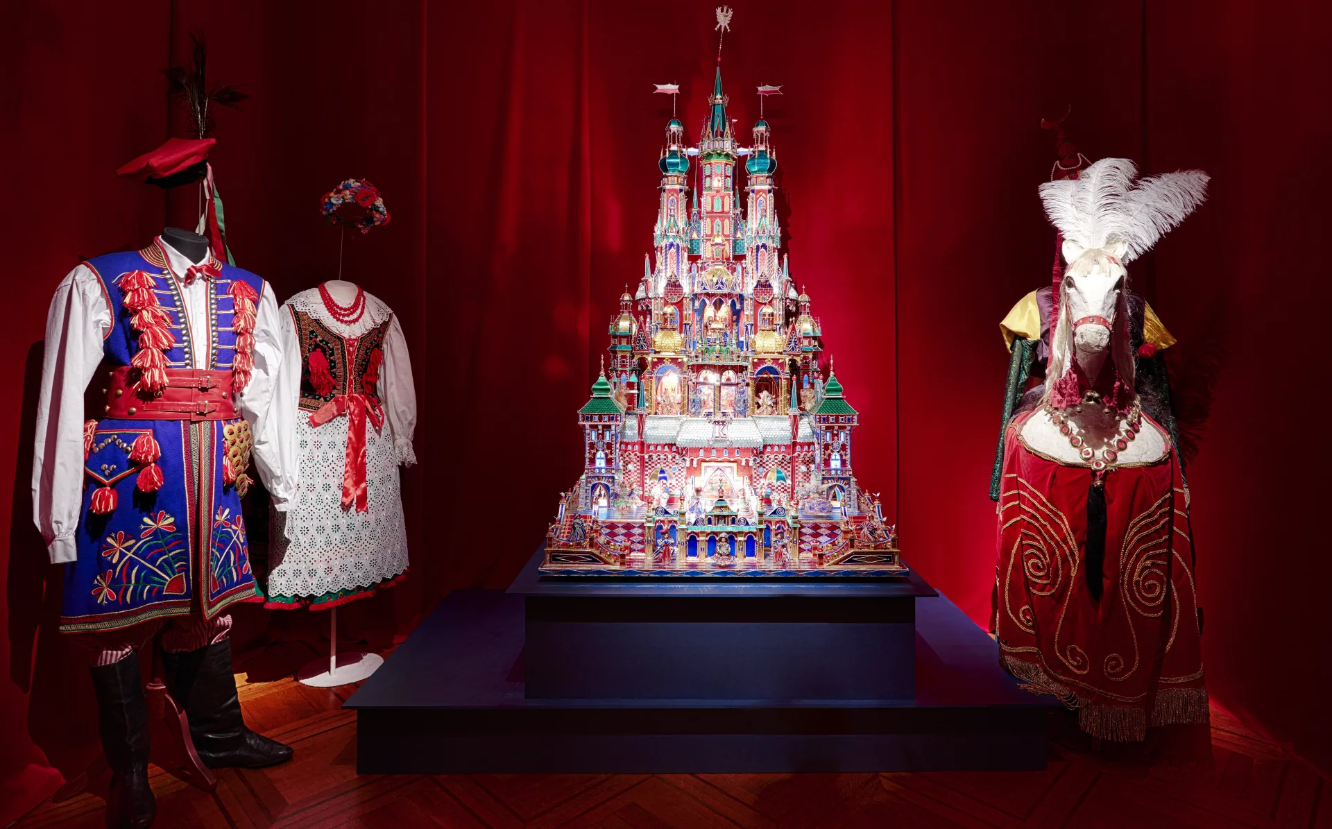 A vibrant museum exhibition with a red backdrop. In the center stands a detailed and glittering nativity scene in the form of a castle, known as a Kraków szopka from Poland. On the left, two traditional Polish folk costumes are displayed on mannequins.