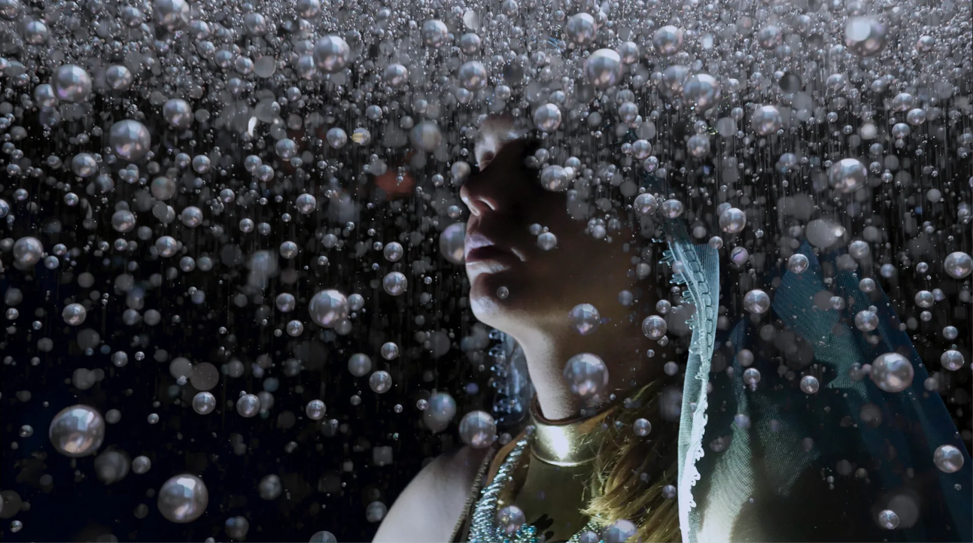 A person is surrounded by countless small, spherical bubbles in a dreamlike, underwater-inspired scene. The face and upper torso are partially visible, adorned with metallic and beaded accessories, including a gold choker and a beaded necklace.
