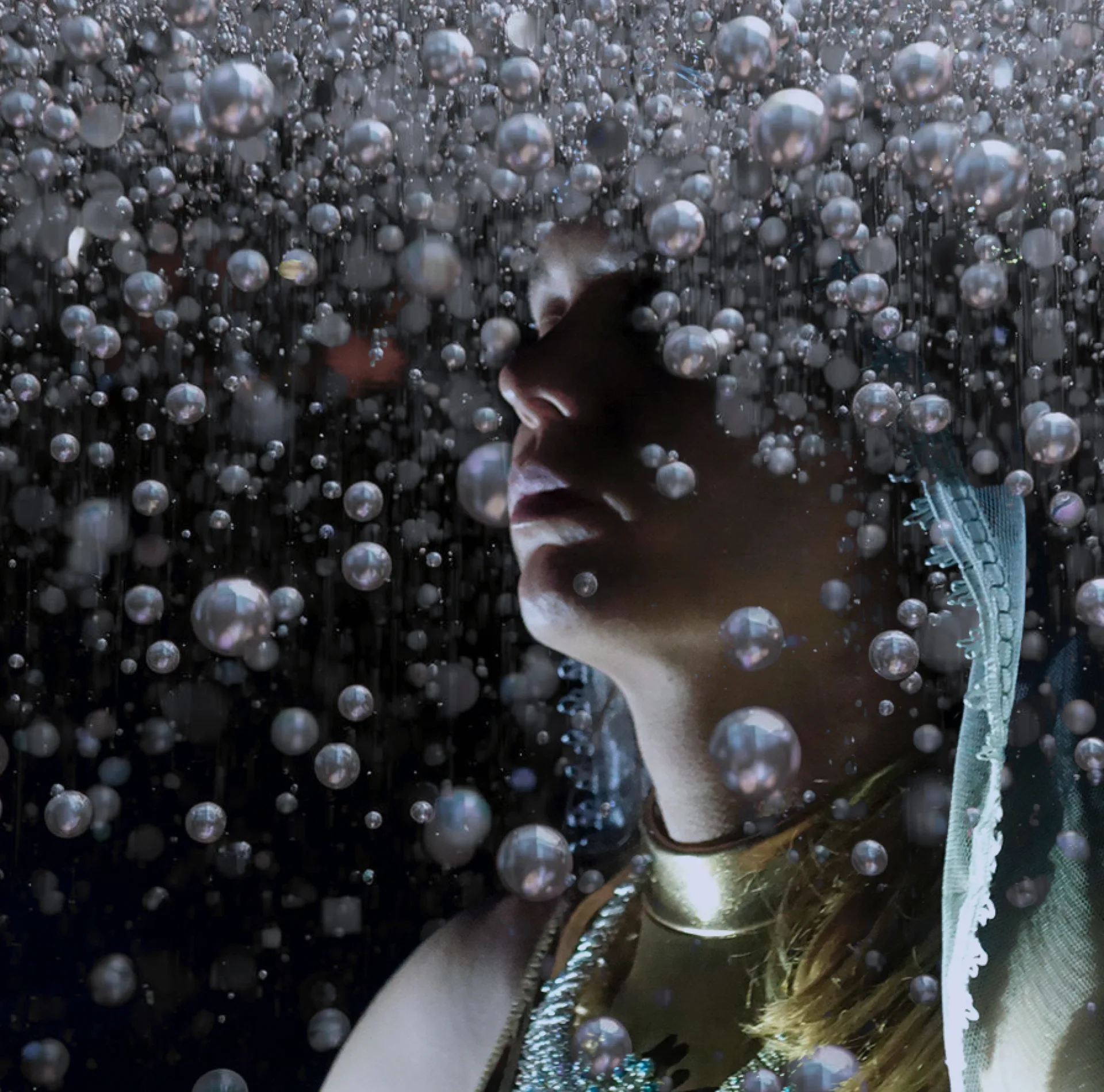 A person is surrounded by countless small, spherical bubbles in a dreamlike, underwater-inspired scene. The face and upper torso are partially visible, adorned with metallic and beaded accessories, including a gold choker and a beaded necklace.