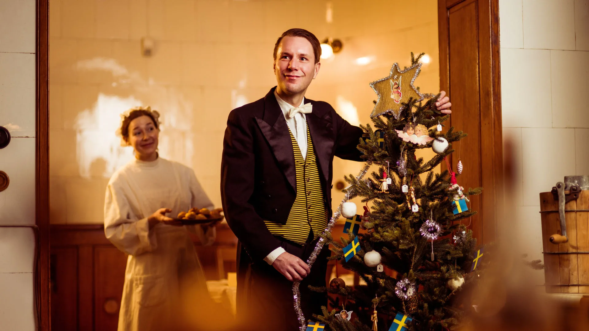 Två personer i historiska kläder firar jul i ett traditionellt inrett rum. Den ena, klädd i svart kostym med gulrandig väst och vit fluga, dekorerar en julgran med svenska flaggor, vita kulor och en blomformad stjärna i toppen. Den andra, i ljus tjänsteklädsel, håller ett fat med nybakade godsaker och ler mot kameran.